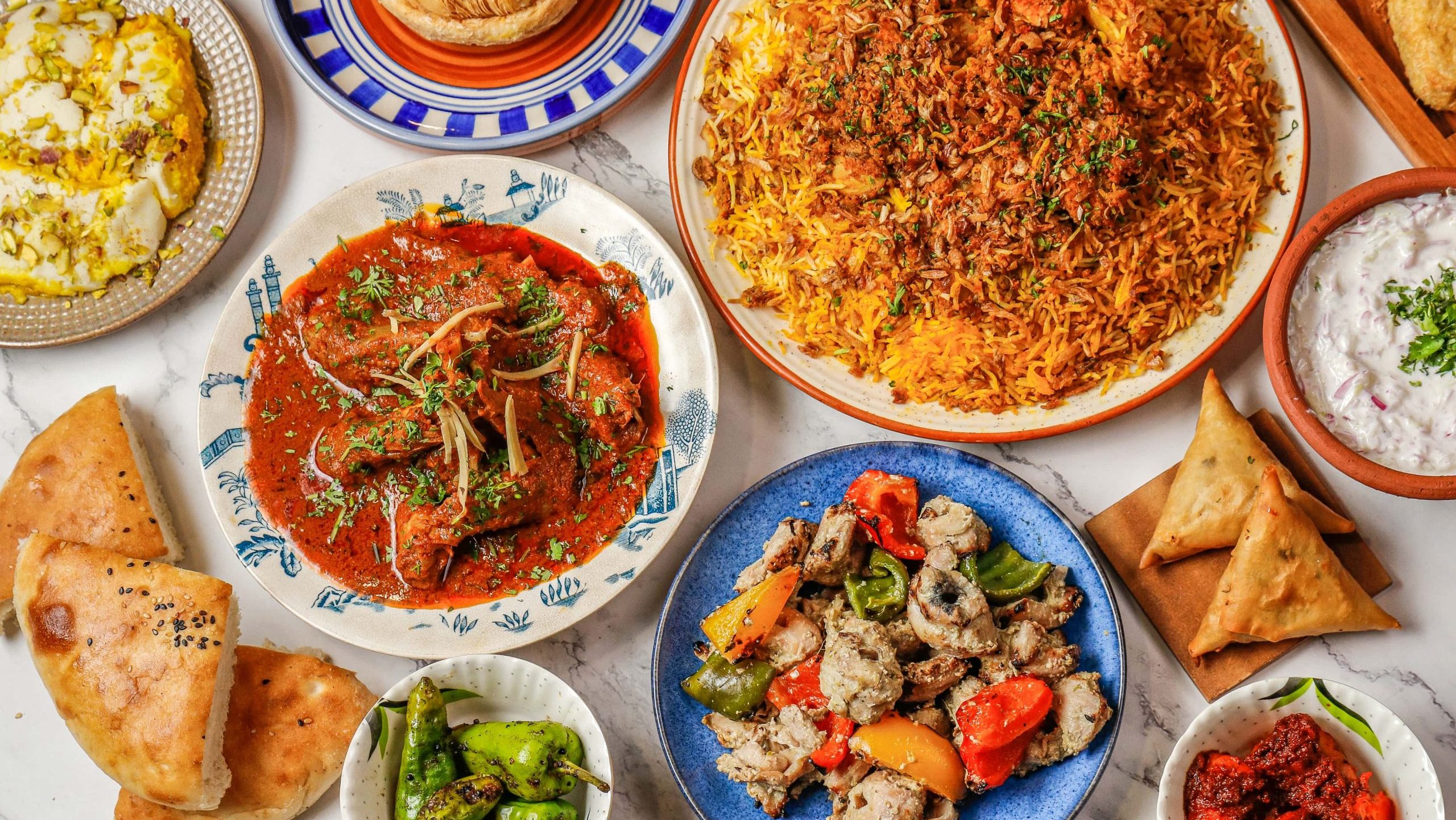 An overhead spread of The Bohri Kitchen dishes: mutton biryani, chicken curry, grilled kebabs, samosas, pav, raita, and condiments on a marble table